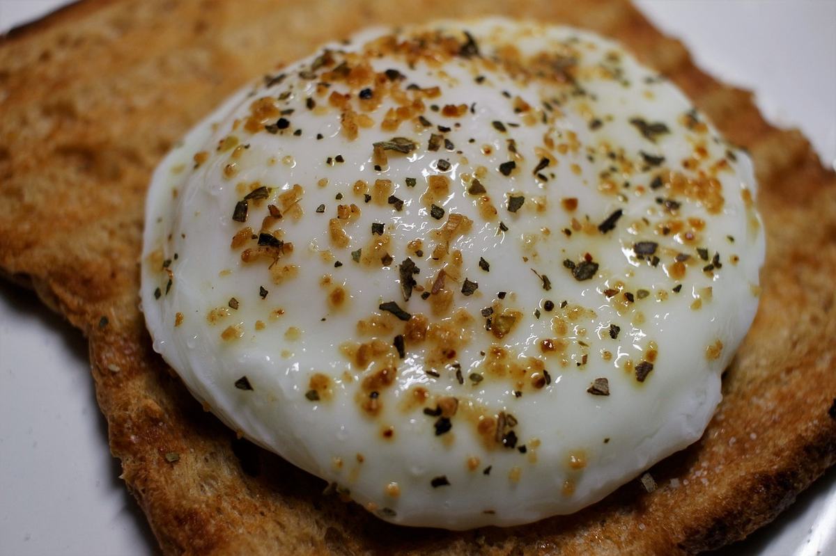 Ein pochiertes Ei mit glattem, geschlossenem Eiweiß liegt auf einer Scheibe geröstetem Brot, bestreut mit Pfeffer und Kräutern.