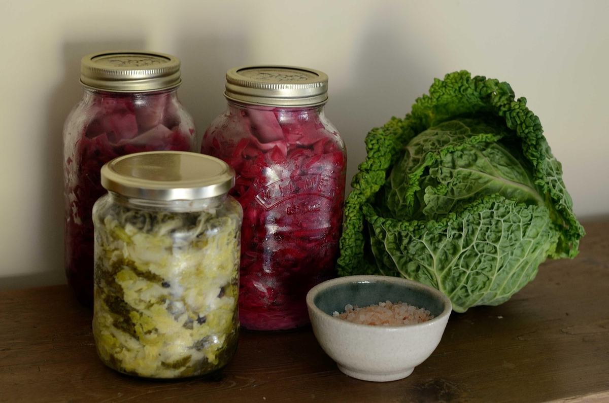 Drei Einmachgläser mit selbst fermentiertem Sauerkraut und Rotkraut neben einem Wirsingkopf und einer Schale mit grobem Salz