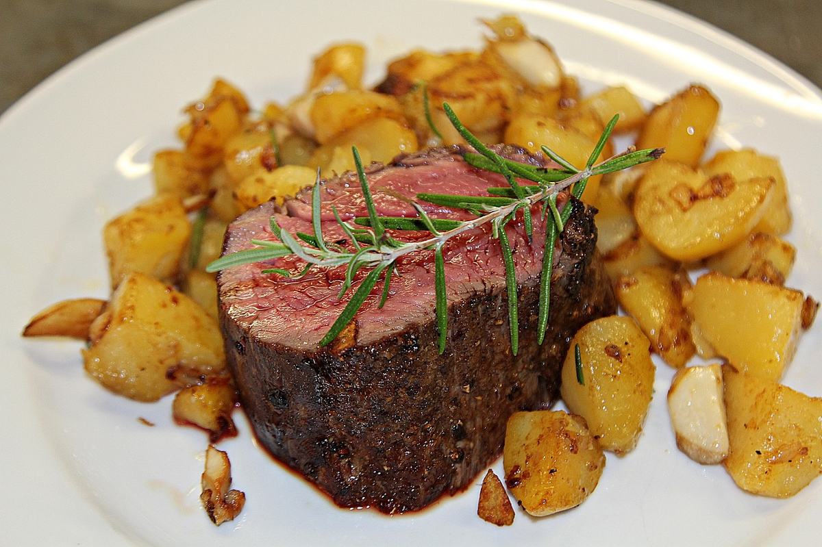 Angeschnittenes Filetsteak mit rosa Kern, Rosmarin und Bratkartoffeln auf einem weißen Teller