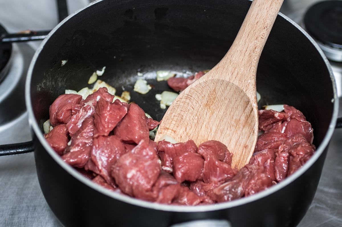 Viele rohe Rindfleischwürfel mit Zwiebeln dicht an dicht in einem schwarzen Topf, ein Holzkochlöffel rührt um