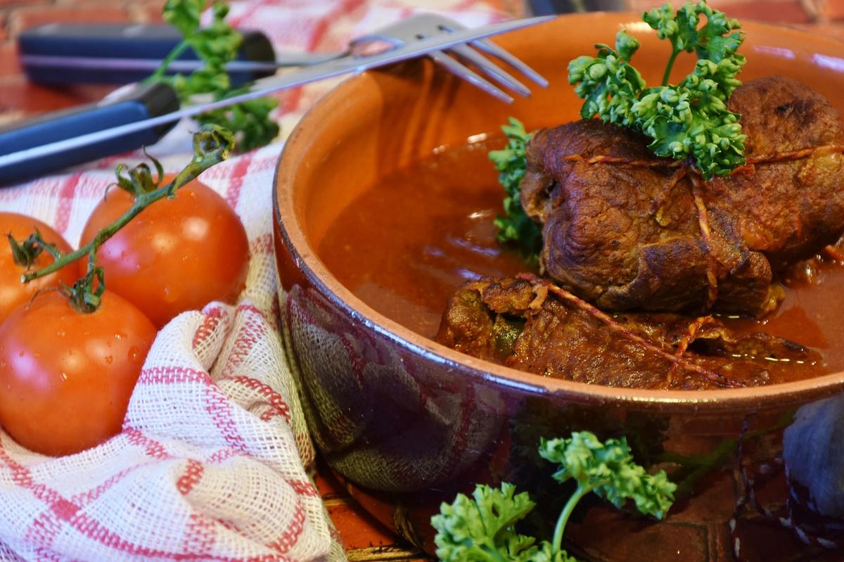 Rinderrouladen in dunkler Schmorsoße in einer Tonschale, daneben Tomaten und Petersilie
