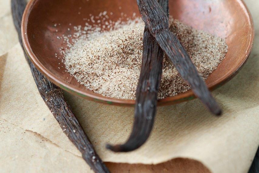 Drei Vanilleschoten liegen auf und neben einer kleinen Kupferschale mit Vanillezucker